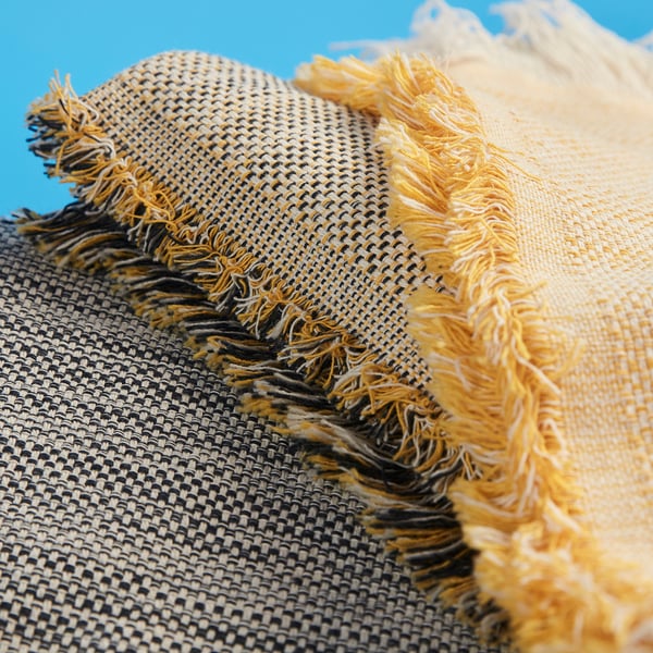 Eine Nahaufnahme eines gewebten, strukturierten Überwurfs mit sichtbaren gelben und weißen Fransen. Der Stoff wirkt weich und warm und zeichnet sich durch ein detailliertes Webmuster aus.