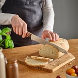 Person, die mit einem gezackten Messer auf einem hölzernen Schneidebrett Brot schneidet.