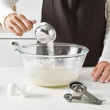 Eine Person gibt mit Hilfe von Messbechern Zutaten in eine Rührschüssel. Die Becher werden auf eine Theke gestellt, bereit für genaue Messungen.