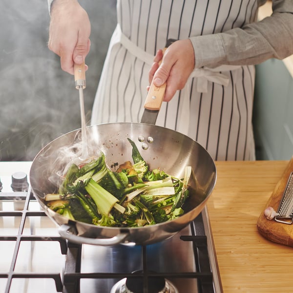 Person brät Gemüse in einem Wok auf dem Herd. Dampf steigt aus dem Wok auf und zeigt an, dass der Garvorgang läuft.