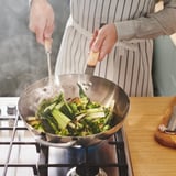 Person brät Gemüse in einem Wok auf dem Herd. Dampf steigt aus dem Wok auf und zeigt an, dass der Garvorgang läuft.
