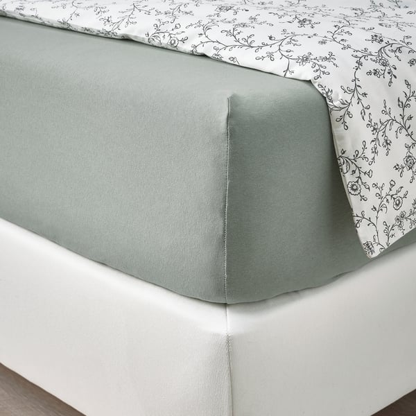 Bett mit grünem Spannbettlaken, gemusterter weißer Bettdecke, auf weißer Matratze. Das Blatt hat dehnbare Ecken.