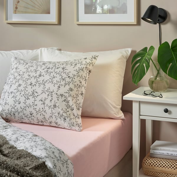 Ein Bett mit weißen VÅRVIAL geblümten Kissenbezügen, einem beigen Kissen und rosa Laken. Daneben steht ein Nachttisch mit einer grünen Pflanze, Büchern und einer Lampe.