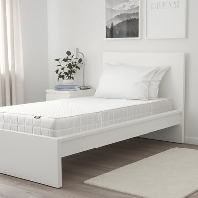 Modernes minimalistisches Schlafzimmer mit weißer VATTAO-Matratze, zwei Kissen, Bett auf dunklem Boden, beigem Teppich, Nachttisch mit Pflanze und Buch, Wandkunst, Vorhängen.