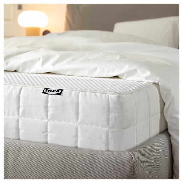 Gemütliches weißes Bett mit einer neuen IKEA-Matratze, weichen Laken und einer stilvollen Lampe.