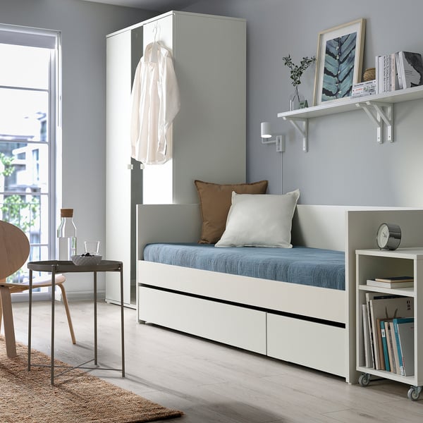 Modernes Schlafzimmer mit weißem Tagesbett, blauen Kissen, Regalen, Stuhl und Bücherregal.