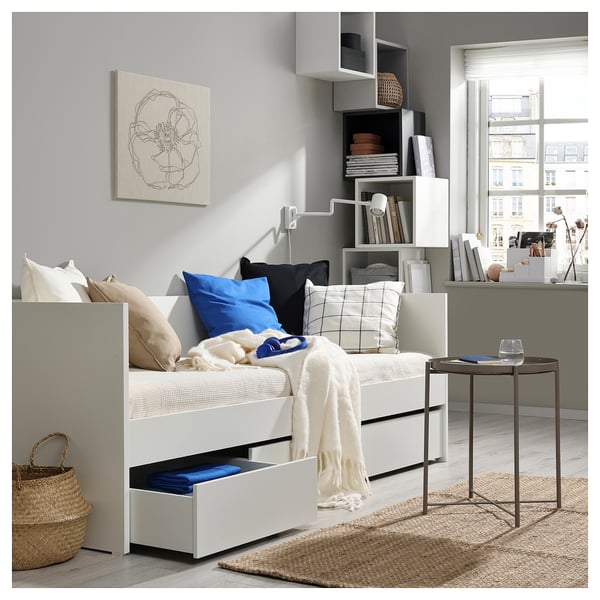 Minimalistisches Schlafzimmer mit weißem Tagesbett und Schubladen. Bunte Kissen, Teppiche und Kunst an der Wand. Schreibtisch mit Stuhl und Blick auf die Stadt aus dem Fenster.