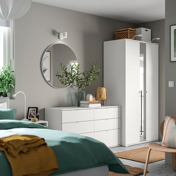 Modernes Schlafzimmer mit blaugrüner Bettwäsche, weißen Möbeln, rundem Spiegel und grüner Pflanze.