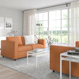 Helles Wohnzimmer mit großen Fenstern. Orange VIMLE Sofa, moderner Couchtisch und Beistelltisch. Weißes Dekor, draußen sichtbares Grün.