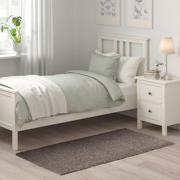 Schlafzimmerszene mit weißem Bett, Kissen und Nachttisch. Grauer Teppich unter dem Bett, Hintergrund mit Pflanzen, Lampen und Wandkunst.