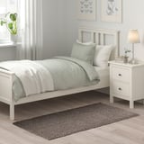 Schlafzimmerszene mit weißem Bett, Kissen und Nachttisch. Grauer Teppich unter dem Bett, Hintergrund mit Pflanzen, Lampen und Wandkunst.