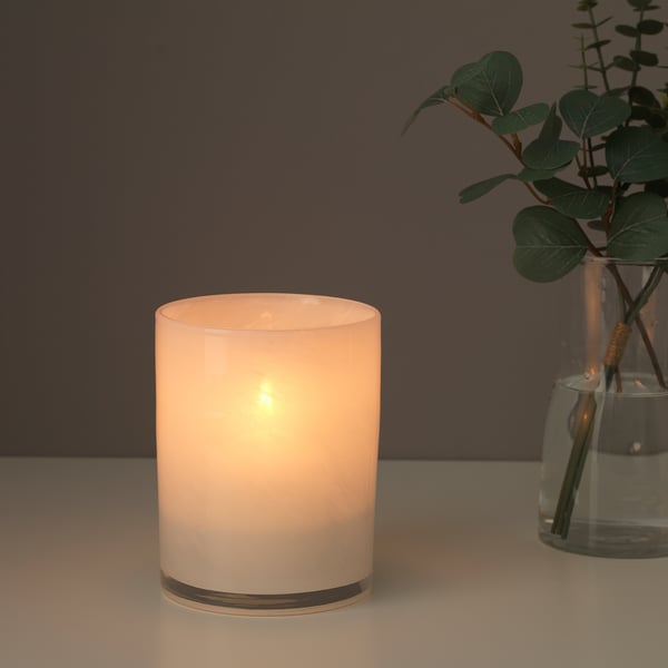Zylindrischer Kerzenhalter aus Glas mit mattiertem, goldenem Sockel. Hält eine brennende Kerze und strahlt warmes, weiches Licht aus. Gruppen mit grüner Pflanze in klarer Vase.