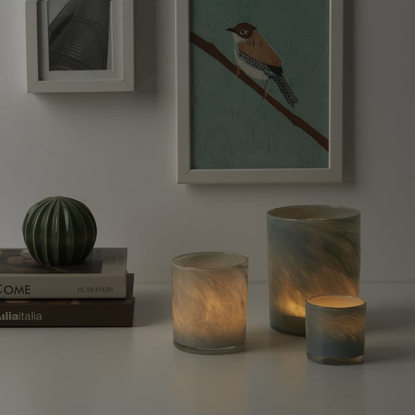 Drei VINDSTILLA-Kerzen in verschiedenen Größen auf dem Tisch mit Büchern, die warm angezündet und leuchten. Kunst im Hintergrund.