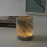 Eine brennende Kerze in einem strukturierten Glashalter sitzt auf einer weißen Oberfläche. Das Design des Halters spiegelt das warme Licht wider und schafft ein gemütliches Ambiente.