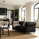Modernes Wohnzimmer mit schwarzem VISKAFORS-Sofa, weißen Vorhängen, Bogenfenstern, Holzboden und schwarzer Küche.