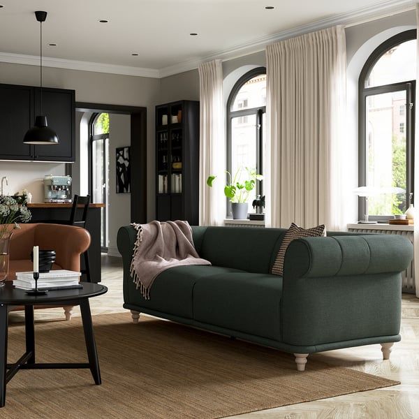 Modernes Wohnzimmer mit grünem VISKAFORS-Sofa, orangefarbenem Sessel, schwarzer Küche und großen Bogenfenstern mit durchsichtigen Vorhängen.