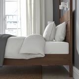 Bett mit hohem Kopfteil aus Holz, weißen Kissen und grauer Decke, neben großem Fenster mit Vorhängen.