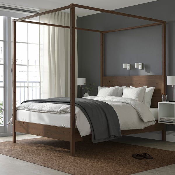 Hölzernes Himmelbett mit braunem Rahmen, weißen Kissen und grauer Bettwäsche. Zentral platziert im Zimmer mit großen Fenstern.