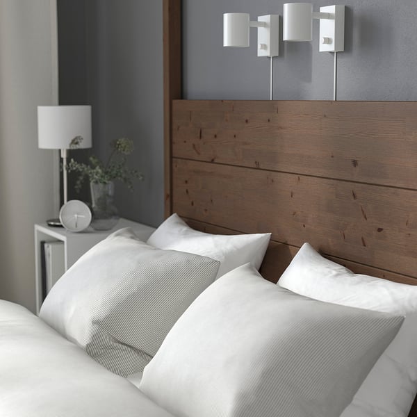 Kopfteilbett aus Holz mit weißen Kissen, Lampe, Pflanze und Uhr auf dem Nachttisch.