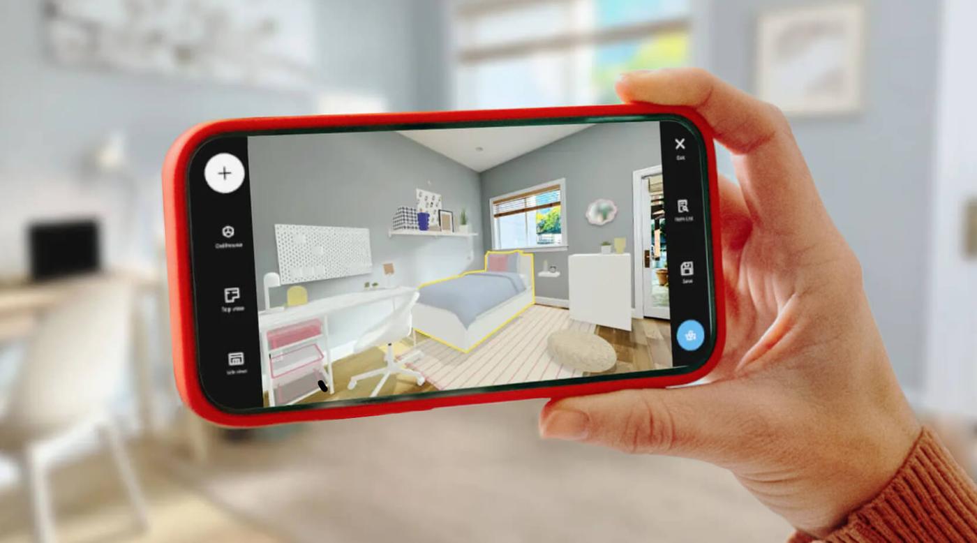 A person holds a smartphone displaying a photographic room scan in IKEA Kreativ, with the real bedroom blurred and partially visible in the background.