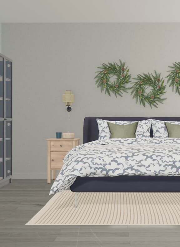 A holiday bedroom design created with IKEA Kreativ, featuring a navy blue bed with a blue and white duvet set, green accent pillows, and a beige area rug. Three decorative wreaths hang above the headboard, framed by matching nightstands and wall lamps.