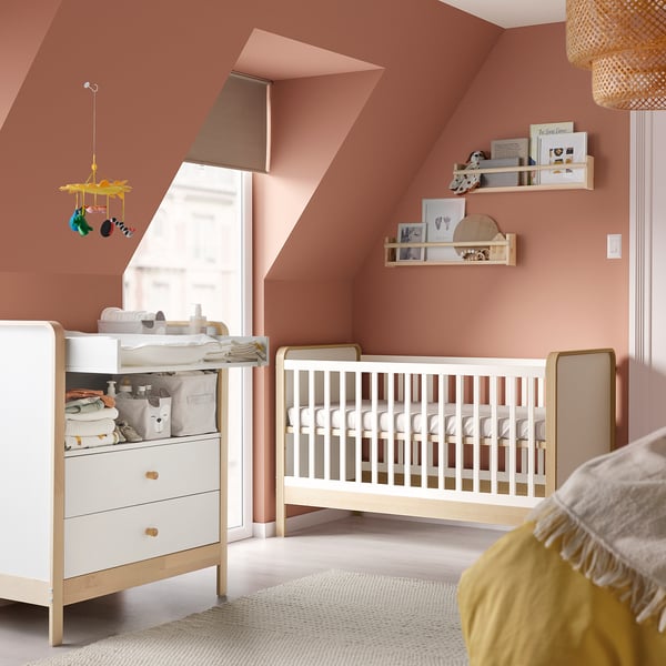 Baby nursery with wooden white crib and dresser, pink walls, yellow hanging mobile, shelves with toys, cozy rug.