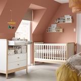 Baby nursery with wooden white crib and dresser, pink walls, yellow hanging mobile, shelves with toys, cozy rug.