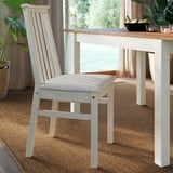 Modern dining chair with a slatted back, cushioned seat, and sturdy pine frame, positioned near a wooden table.
