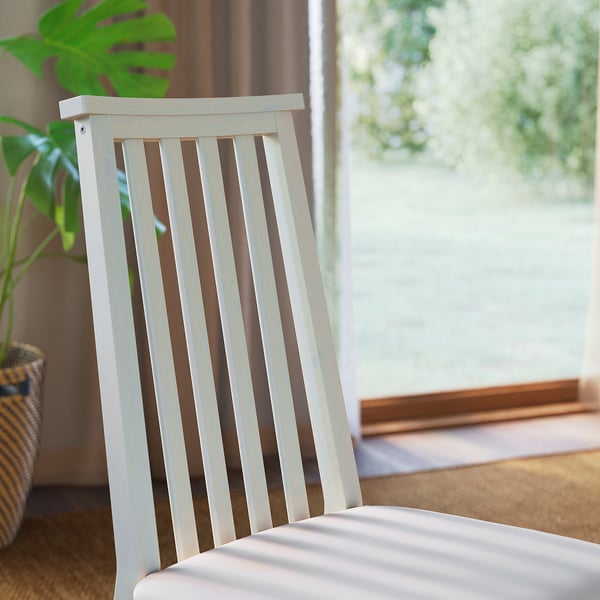 Cozy indoor scene: white slatted chair by window, lush plant, brown curtain, light wooden floor.