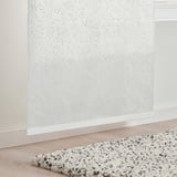 Sheer curtain with leaf pattern allows filtered light. Positioned near a speckled rug on a wooden floor.