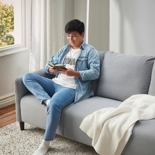 A young person sits on a gray ANGERSBY sofa, reading a book. Soft, comfortable, and practical with side pockets.