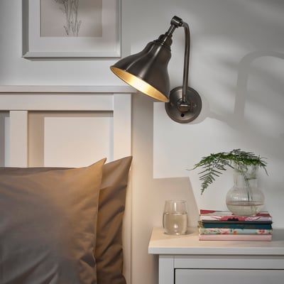 Black ANKARSPEL wall lamp with brass interior above white bedside table. Illuminates table with books, water, and plant. Cozy bedroom decor.