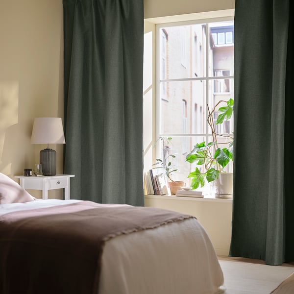 Green curtains in bedroom, drawn to side, framing window with building view. Bed with beige bedding, plants, books, and lamp on nightstand.