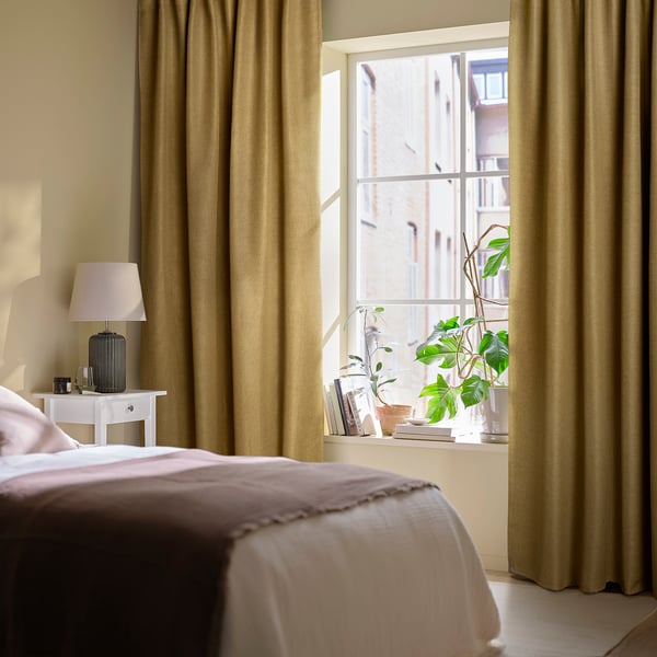 Bedroom with ANNAKAJSA olive curtains, white bed, lamp. Window shows plants, books.