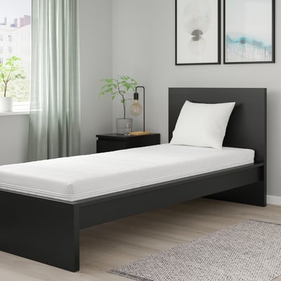 Minimalist bedroom with black ÅNNELAND twin bed, white pillow, wooden floor, light gray walls, green curtains, and a nightstand with lamp and plant.