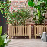Two wooden ASKHOLMEN plant boxes against a brick wall, each with lush green plants, positioned side-by-side, larger than a nearby watering can.
