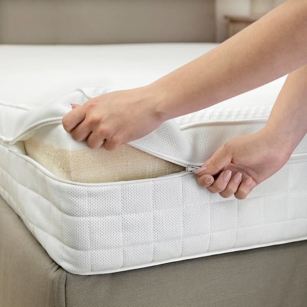 A person zips a white mattress protector onto a foam mattress, showcasing its practical design for easy use and maintenance.