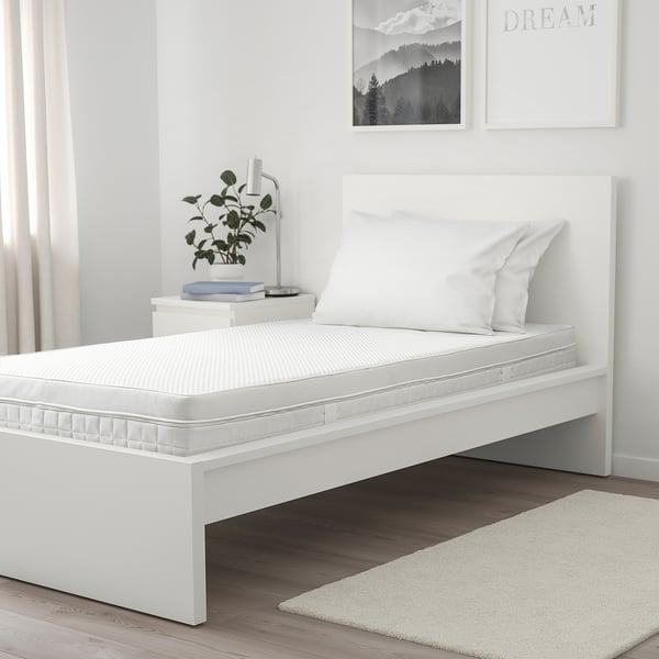 Minimalist bedroom: white ÅVABERG bed, rustic floor, wall art. Bed is small, with two pillows, plant, books, and lamp on nightstand.