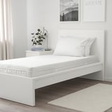 Minimalist bedroom: white ÅVABERG bed, rustic floor, wall art. Bed is small, with two pillows, plant, books, and lamp on nightstand.