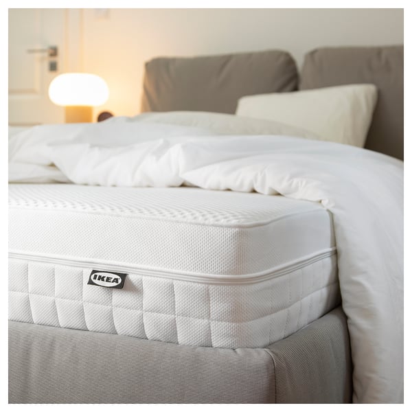 Close-up of a white IKEA mattress with comfort zones and handles, on a bed in a cozy room.