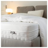 Close-up of a white IKEA mattress with comfort zones and handles, on a bed in a cozy room.