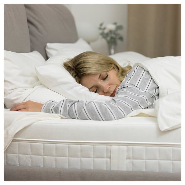 A woman sleeps peacefully on a white ÅVABERG mattress with a grey striped shirt. The room is serene with white linens and a flower vase nearby.