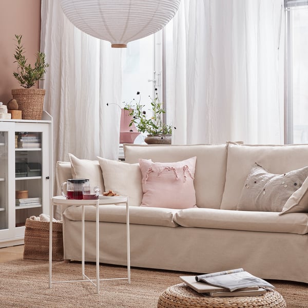 Cozy living room with beige BACKSÄLEN sofa, pink pillows, white curtains, and a side table with drinks.