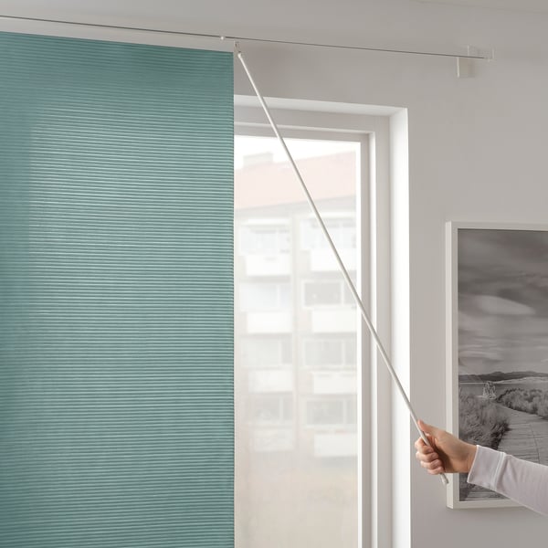 Person opening teal-colored roller blind to let in light, mounted next to framed landscape photo.