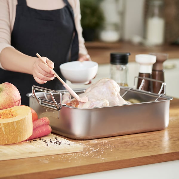 Person preparing meat in a stainless steel roasting tin with rack, surrounded by vegetables and seasonings.