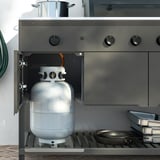 Gas grill cabinet with propane tank, knobs, and skillet.