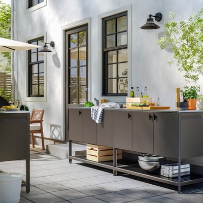 Outdoor kitchen with grey BÅTSKÄR unit, sink, items, chair, plant, large windows.