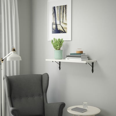Gray armchair next to small white table. Wall shelf with green plant and books. Art print above.