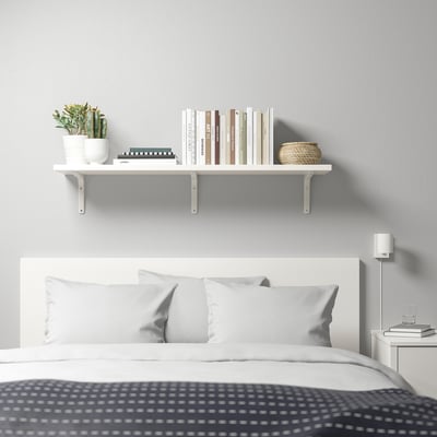 Minimalist bedroom with BERGSHULT shelf holding books, plant, basket. White bed, grey headboard, bedside lamp.