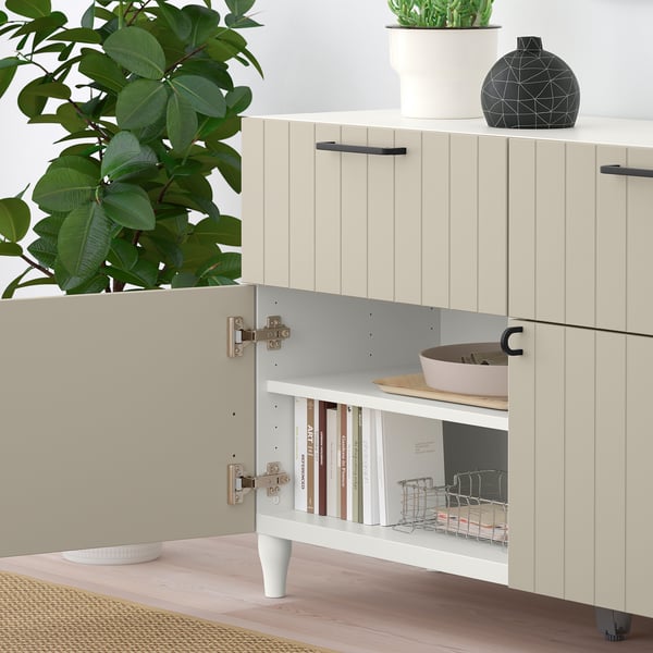 Open BESTÅ sideboard with adjustable shelves, displaying books and kitchen items, against a wall with plants nearby.